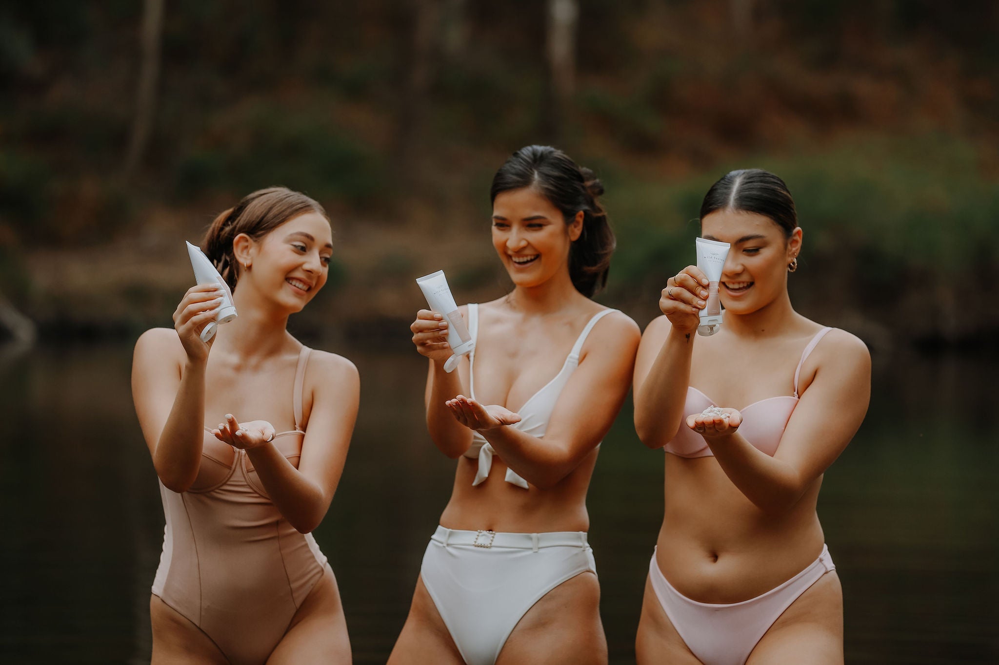 three models holding a tube of wild fusion skincare facial polish. they are smiling and squirting product into their hands. dwellingup forest and lake in background